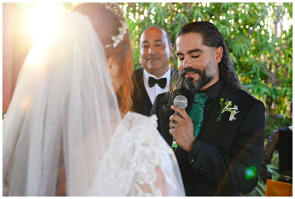 Groom in black tuxedo reads vows into microphone during outdoor garden wedding ceremony.