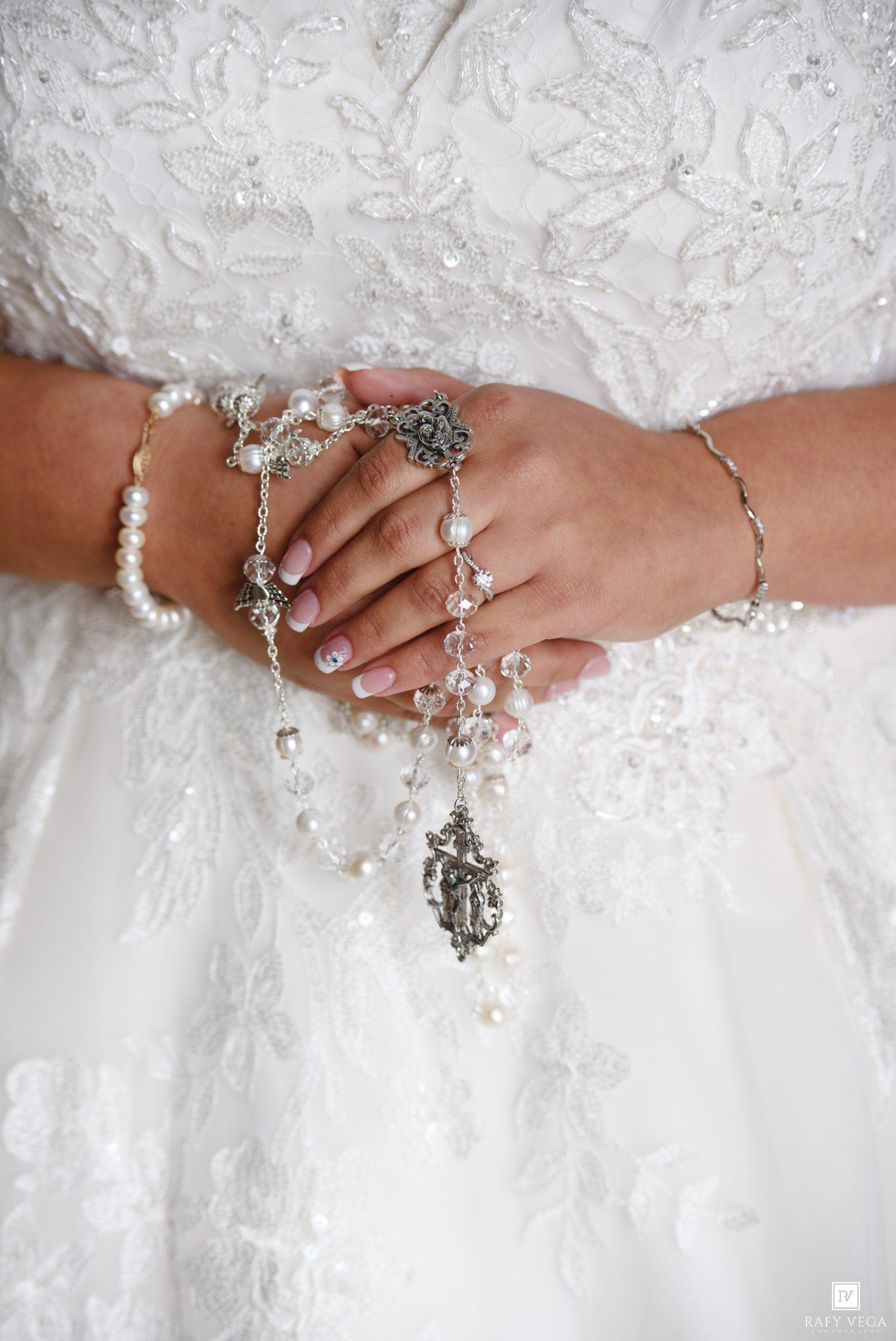 Close-up of bride's hands holding a pearl and crystal rosary, wearing delicate pearl bracelets and white lace wedding gown.