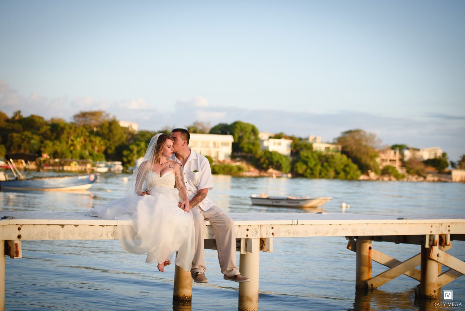 Bride and groom share a romantic kiss on a wooden dock over calm blue waters at sunset.