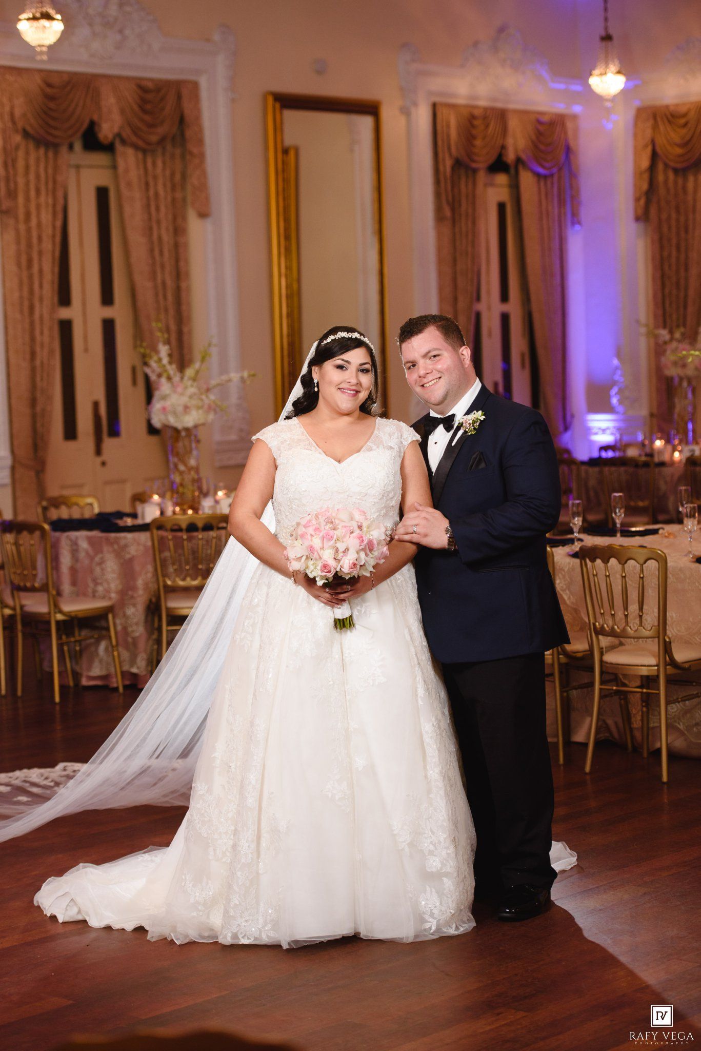 Bride in white lace gown holding pink bouquet poses with groom in navy tuxedo on dance floor at elegant wedding reception.