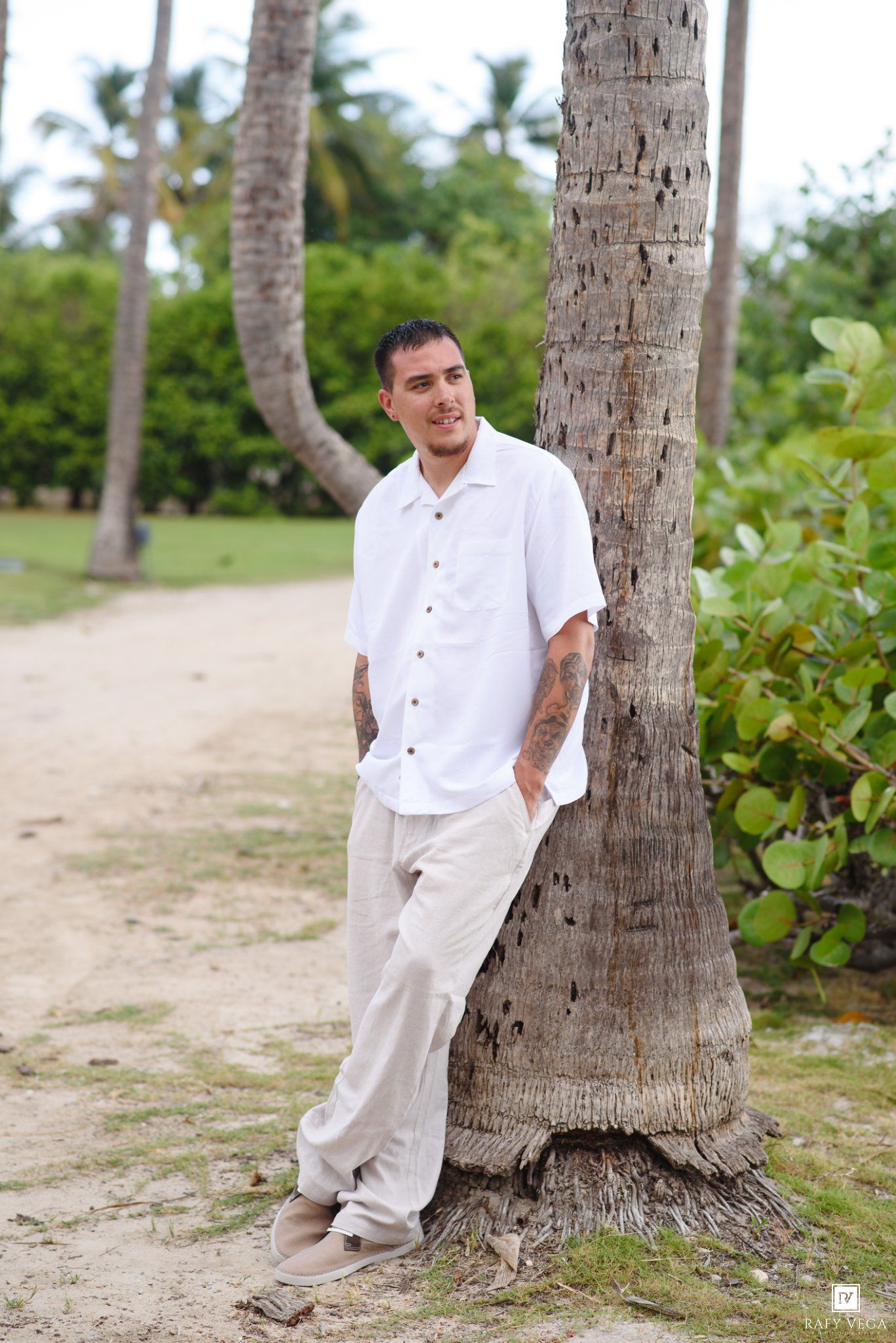 Smiling man with tattoos in white shirt leaning against a palm tree on a sandy tropical path.