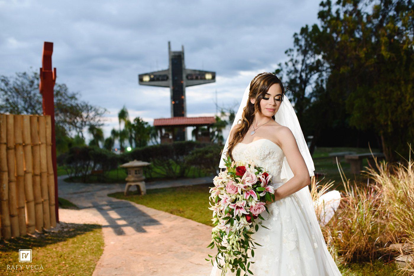 Elegant bride in white gown holds cascading floral bouquet with a cross monument in the background.