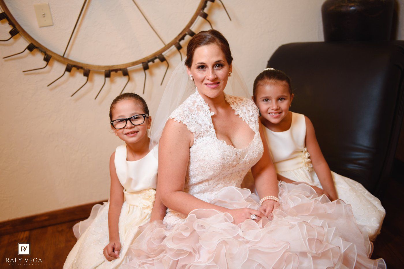 Bride in white lace wedding gown poses with two young flower girls in ivory dresses on wedding day.
