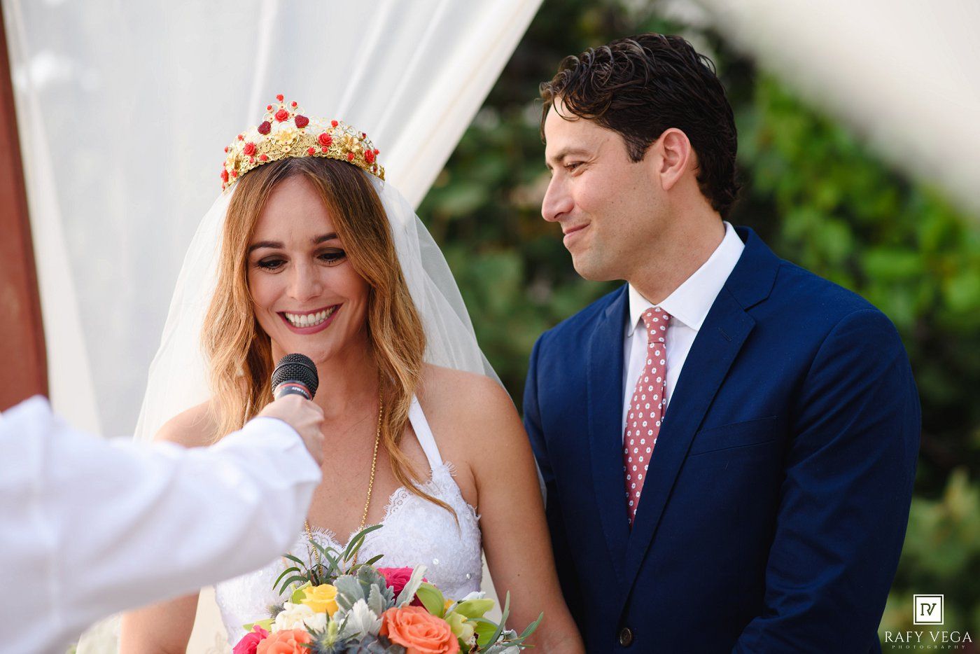 Smiling bride in white gown and red crown holds bouquet beside groom in blue suit at outdoor wedding ceremony.