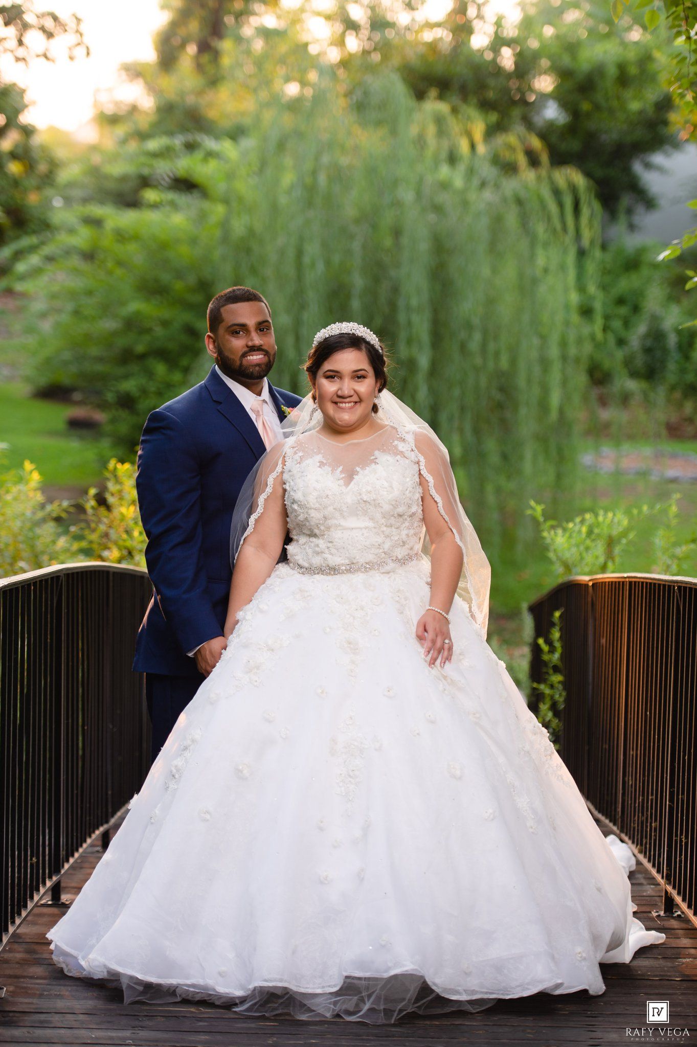 Bride in a ball gown and groom in navy suit pose on a wooden bridge in a garden.