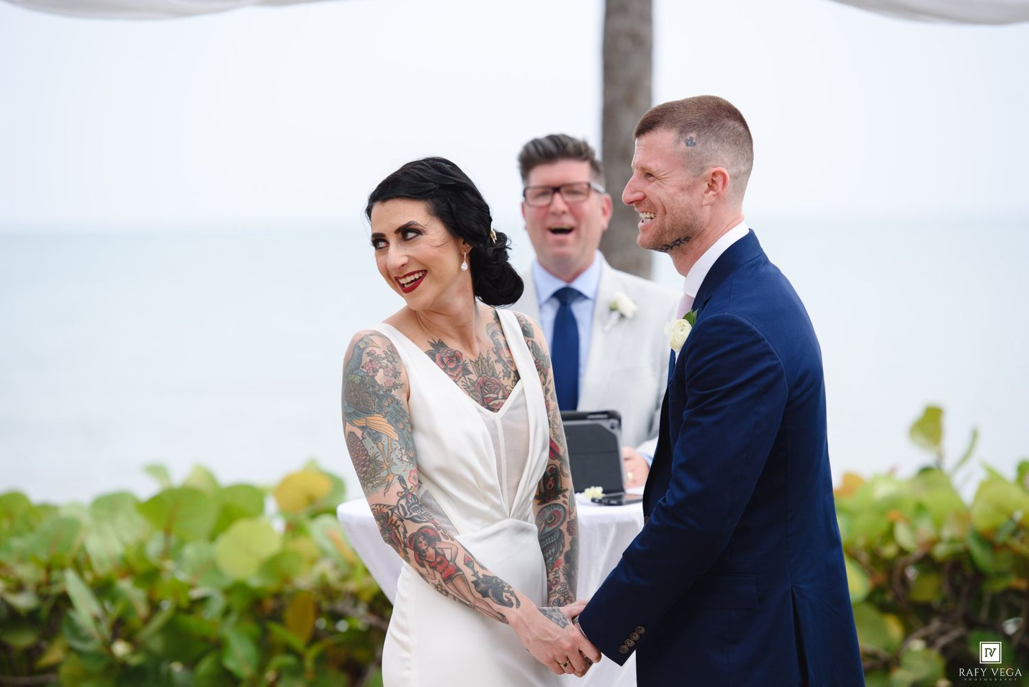 Tattooed bride and groom hold hands and smile during outdoor beach wedding ceremony with officiant behind them.