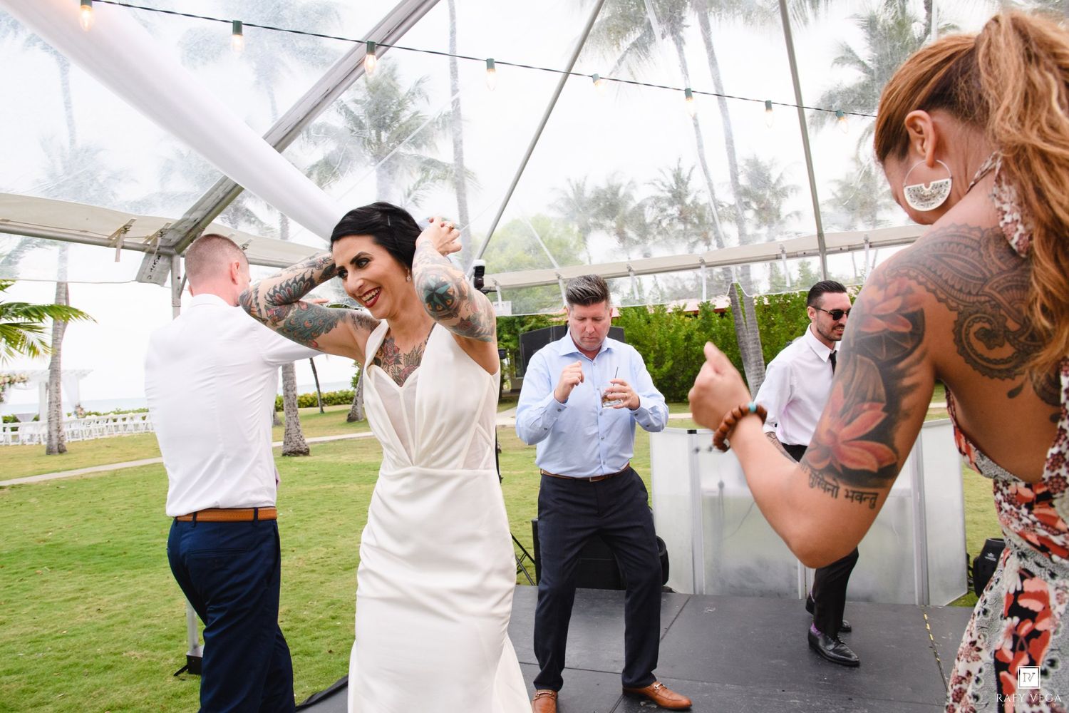 Tattooed bride in white dress dancing joyfully with guests under a tent at an outdoor tropical wedding reception.