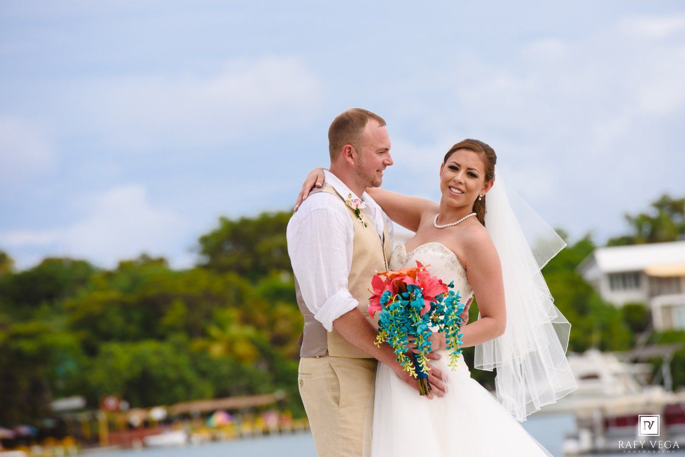 Bride and groom pose outdoors near a marina, bride holding colorful coral and teal bouquet in tropical wedding setting.