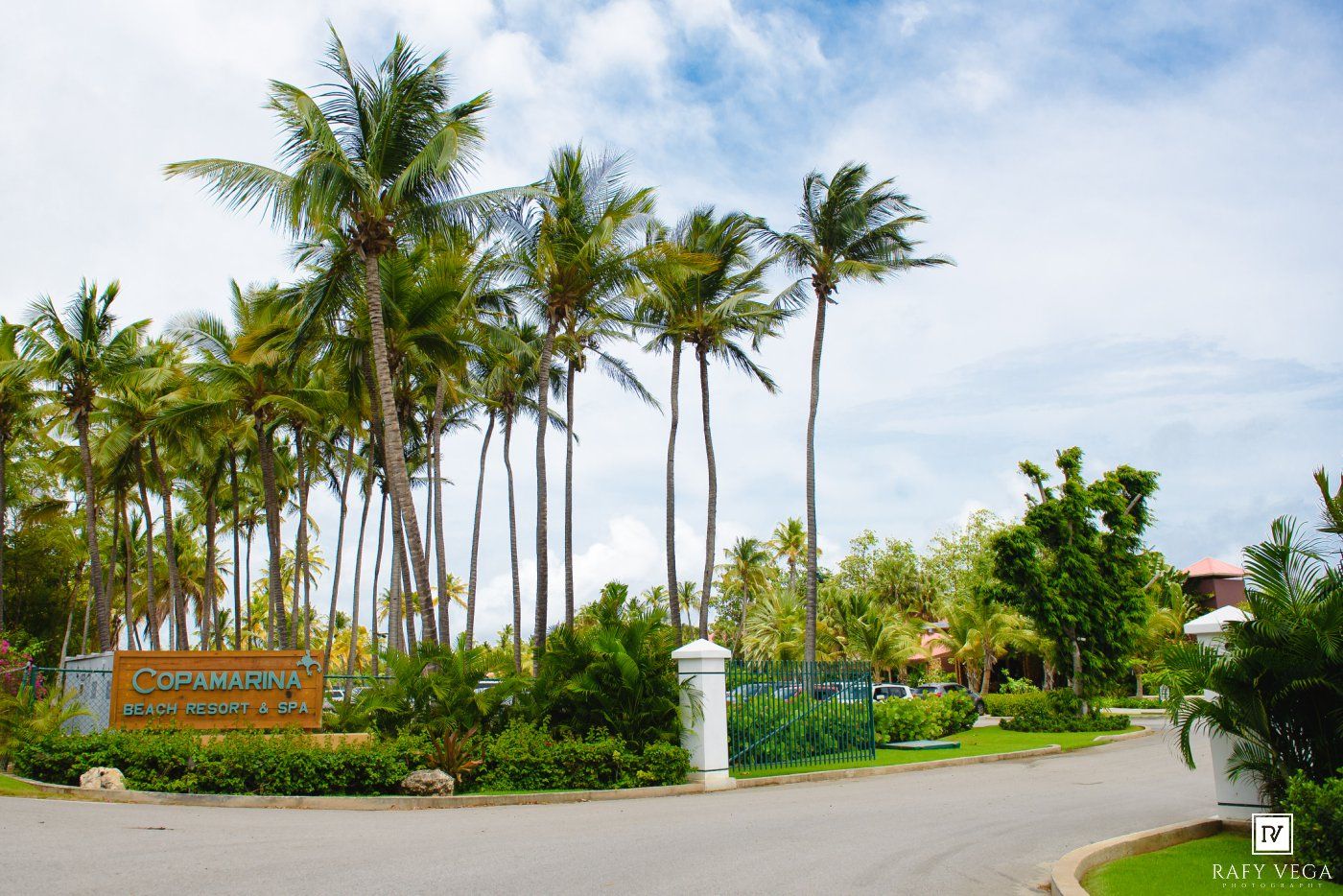Tropical resort entrance with tall palm trees, lush greenery, and a white pillar gate under a partly cloudy sky.