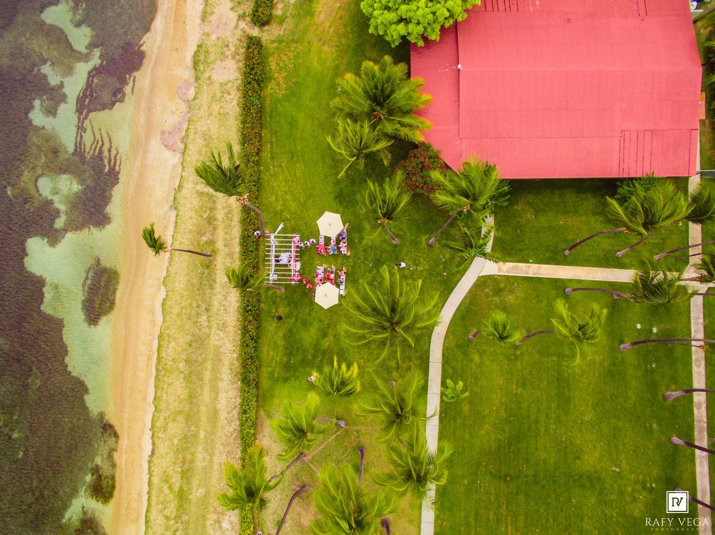 Aerial drone view of tropical beach wedding ceremony beside red-roofed building and palm trees.