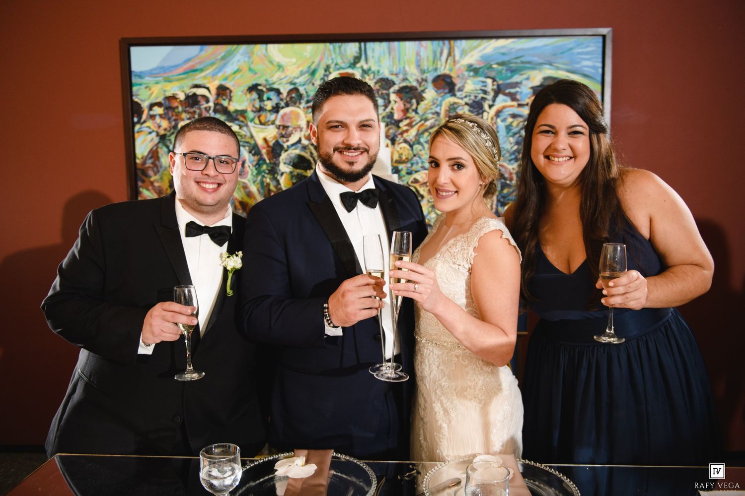 Wedding party of four smiling and toasting with champagne glasses in front of colorful artwork at a reception venue.