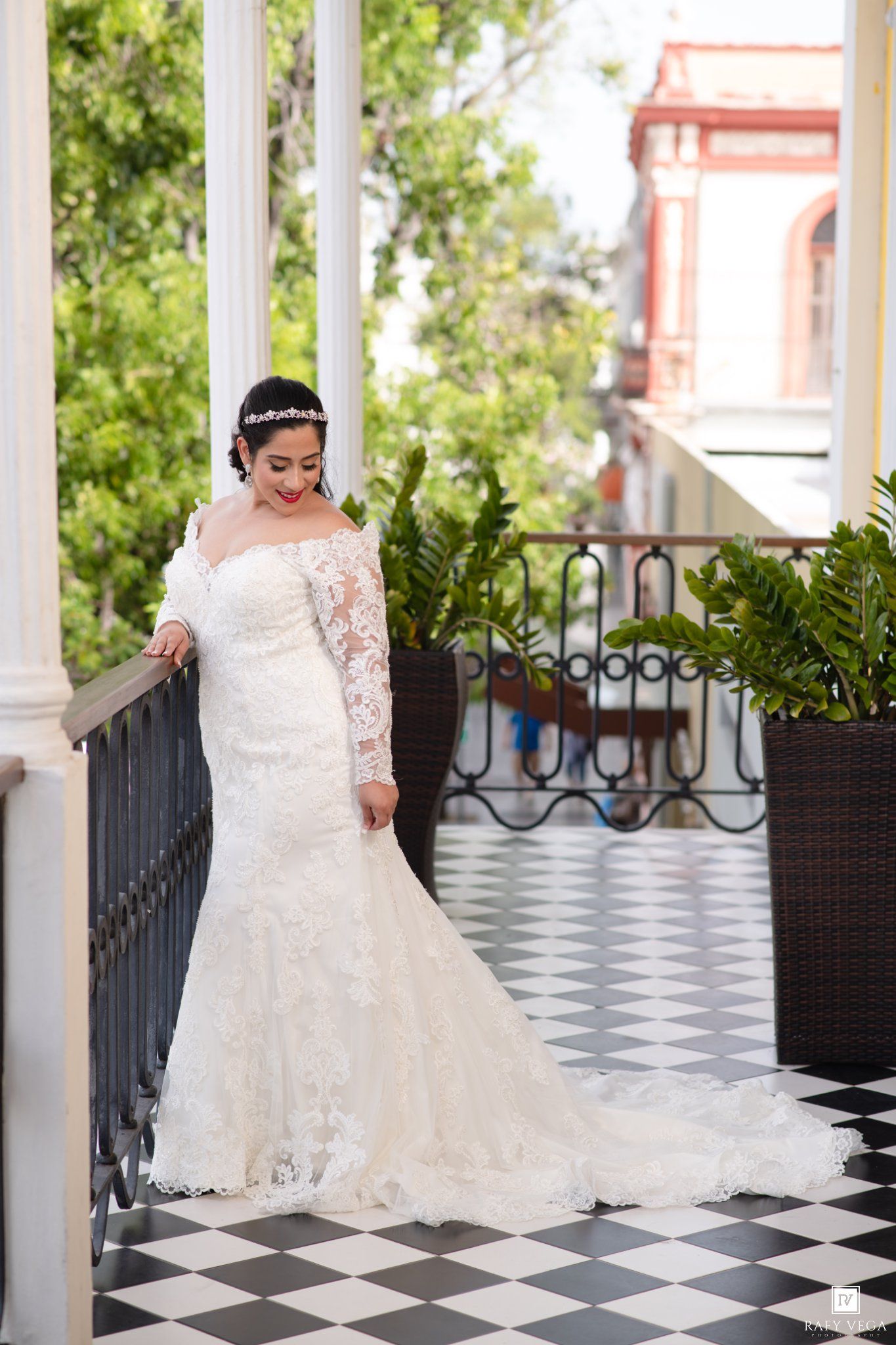 Elegant bride in a long-sleeve lace mermaid wedding gown standing on a checkered balcony with lush greenery backdrop.