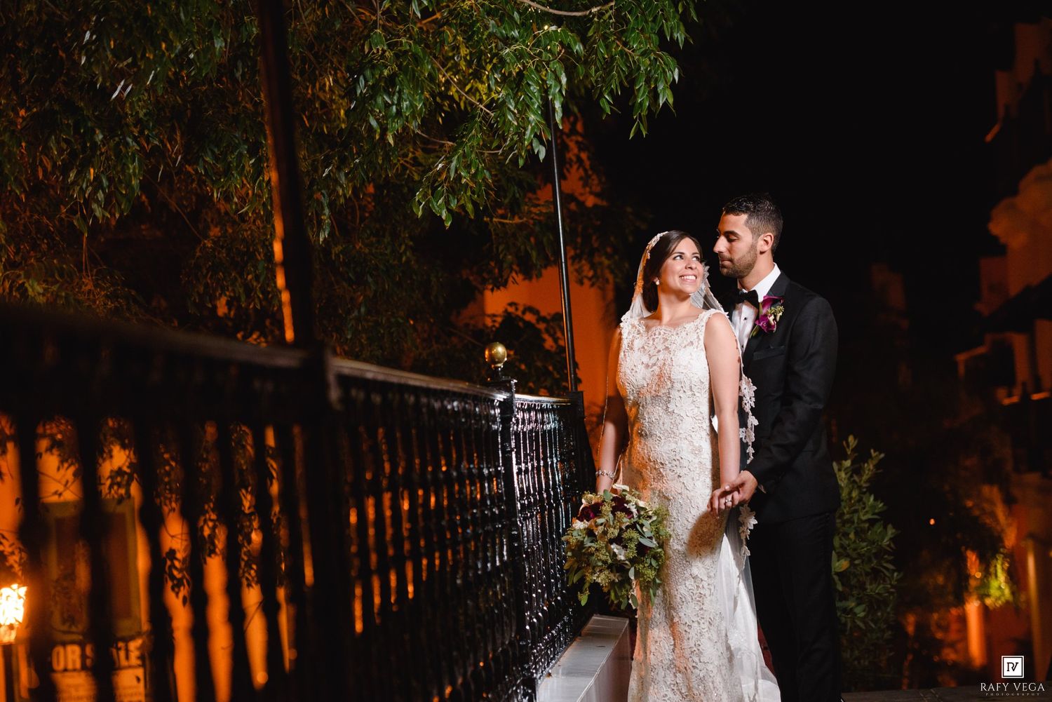 Bride and groom share a romantic moment on an ornate balcony at night, surrounded by warm golden lights and lush greenery.
