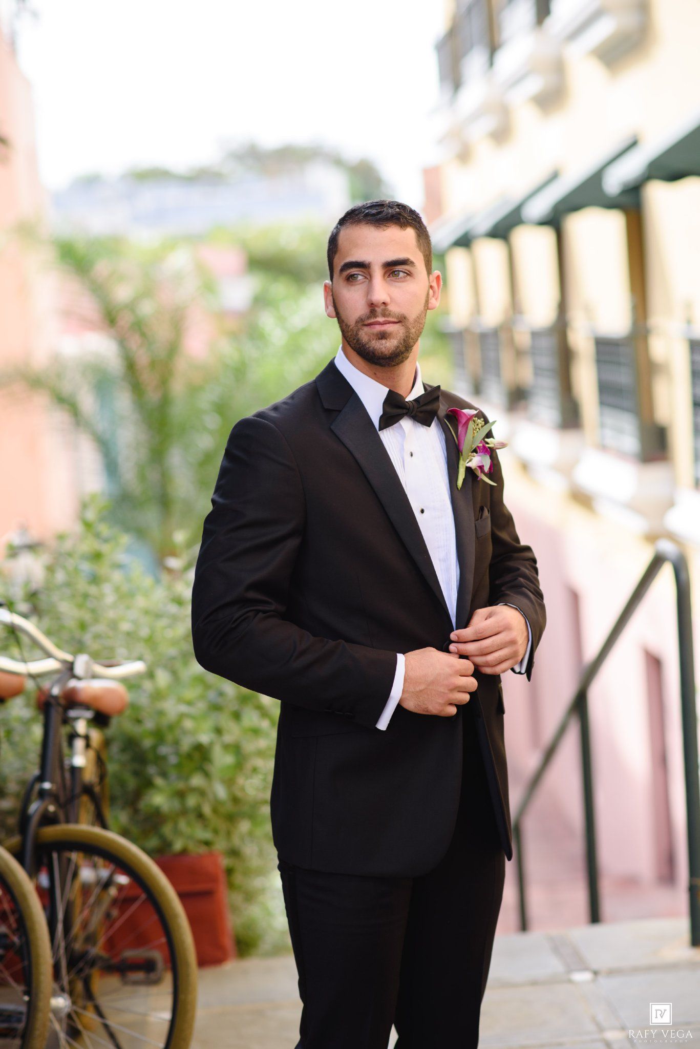 Groom in black tuxedo with bow tie and boutonniere poses outdoors on a city street on his wedding day.