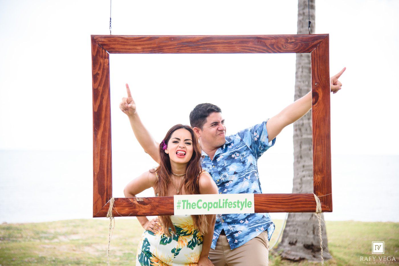 Couple posing in a wooden photo frame prop with #TheCopaLifestyle sign at a tropical outdoor event.