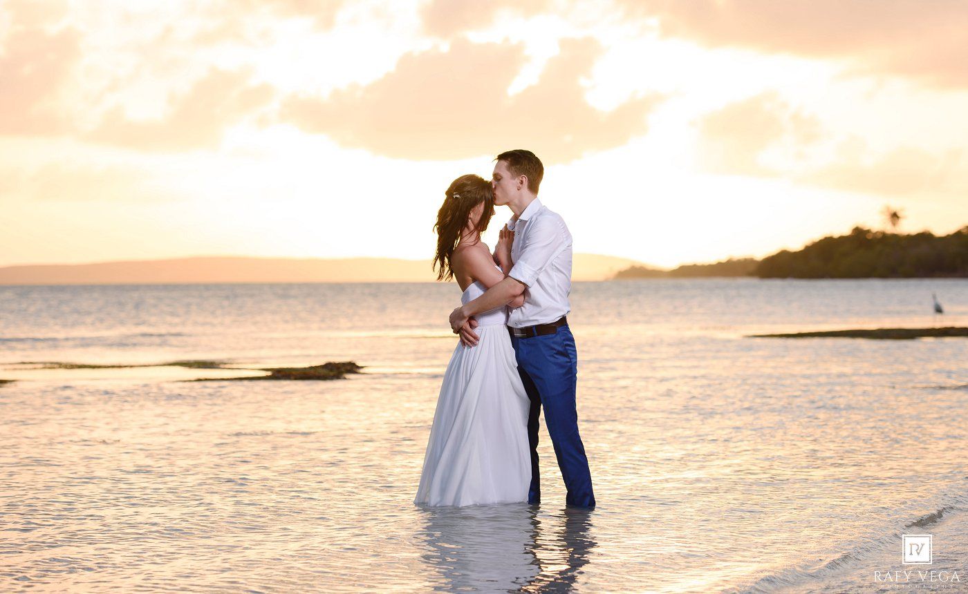 Couple sharing a romantic kiss on a beach at sunset, bride in white gown and groom in blue suit standing in shallow water.