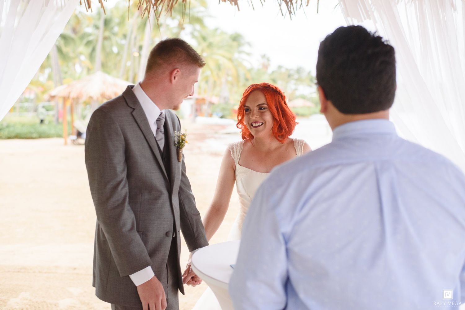 Bride with red hair and groom in gray suit hold hands during outdoor beach wedding ceremony with officiant.