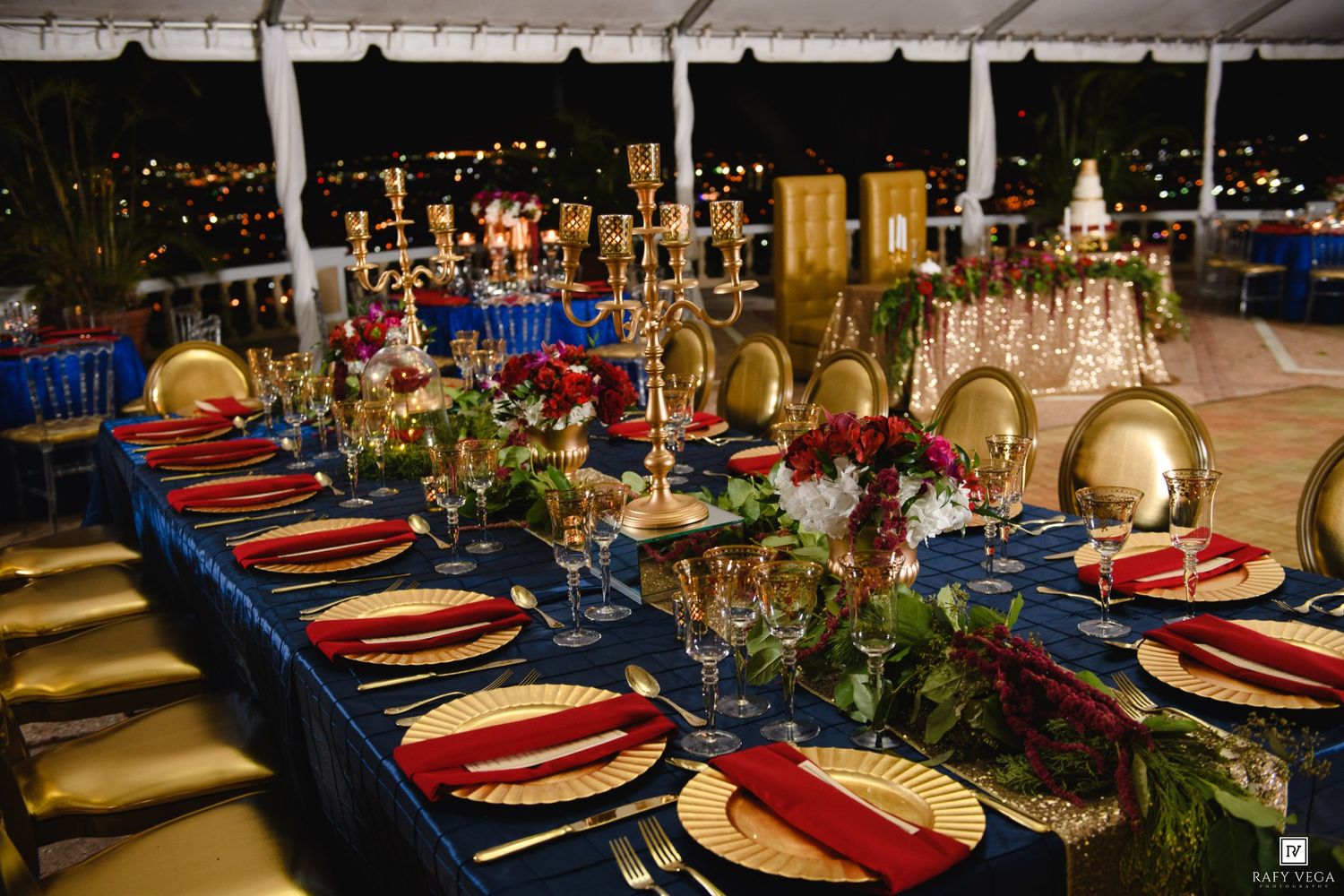 Elegant Beauty and the Beast wedding reception with navy blue tablecloth, gold chargers, red napkins, and candelabras under a tent.