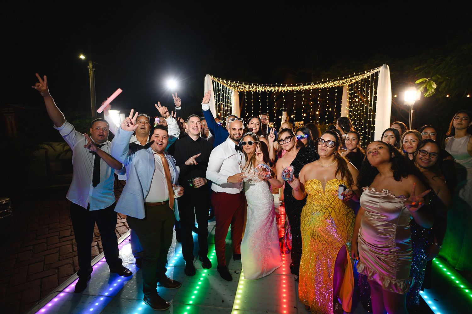 Wedding party celebrating on colorful LED dance floor at night with string lights backdrop.