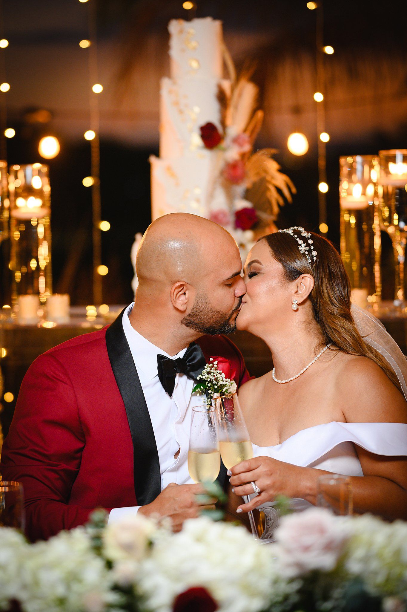 Newlyweds sharing a romantic kiss at wedding reception with champagne and glowing cake backdrop.