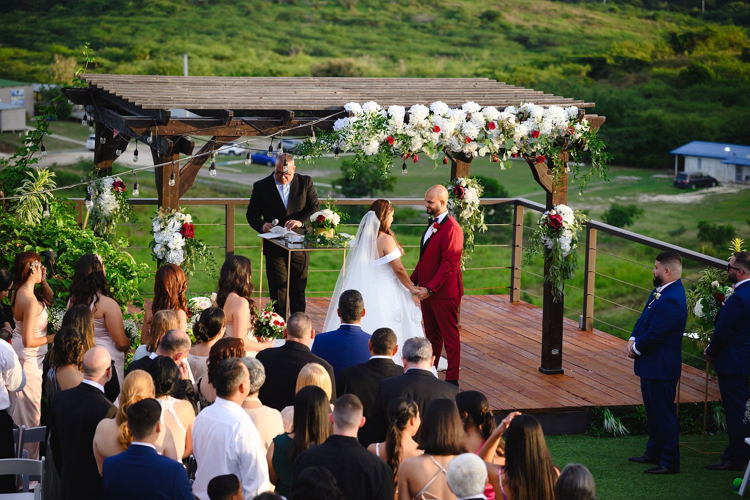Outdoor wedding ceremony on a wooden deck with floral arch, bride and groom at altar, officiant, and seated guests.