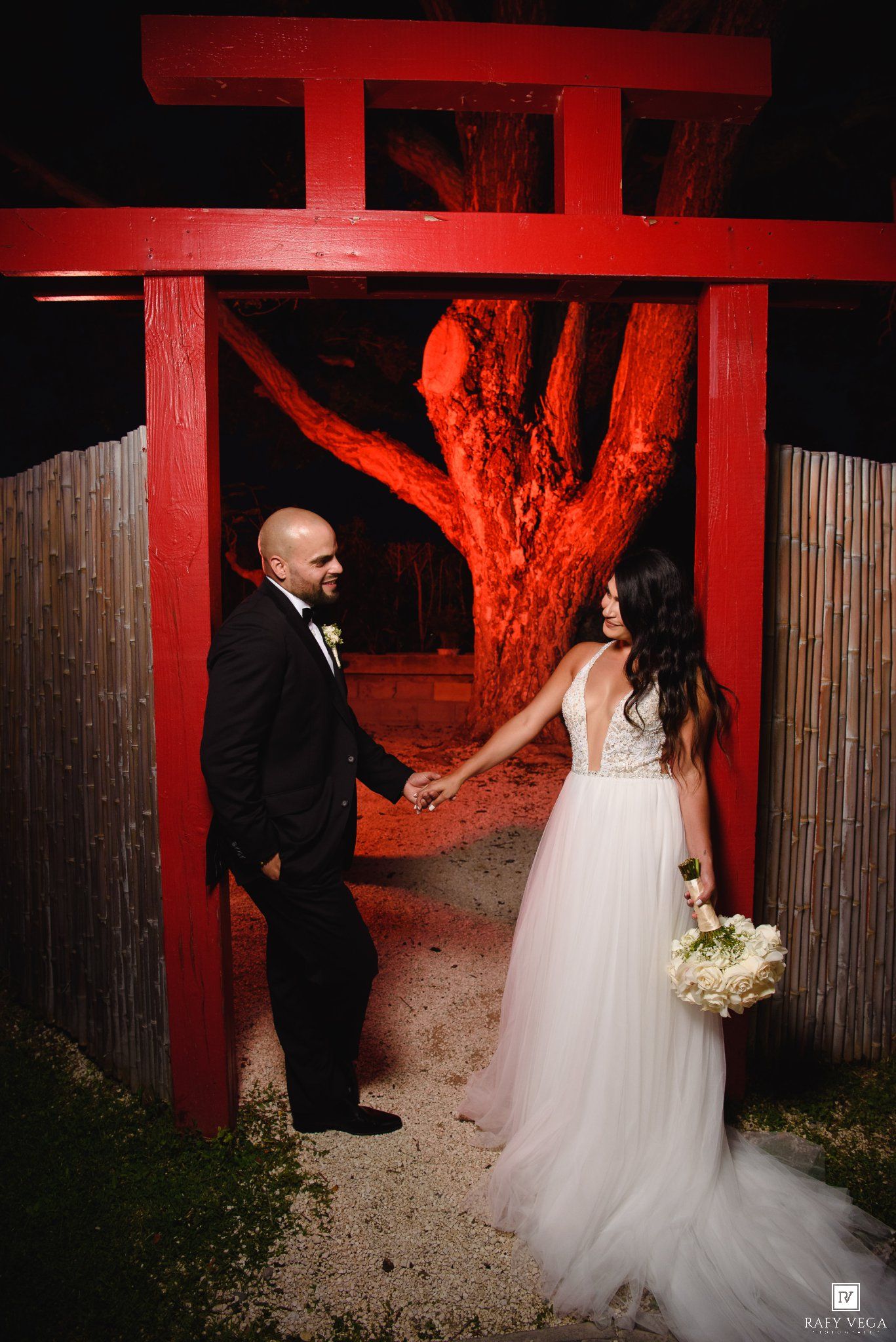 Bride and groom holding hands under a red torii gate at night with a dramatically lit tree behind them.