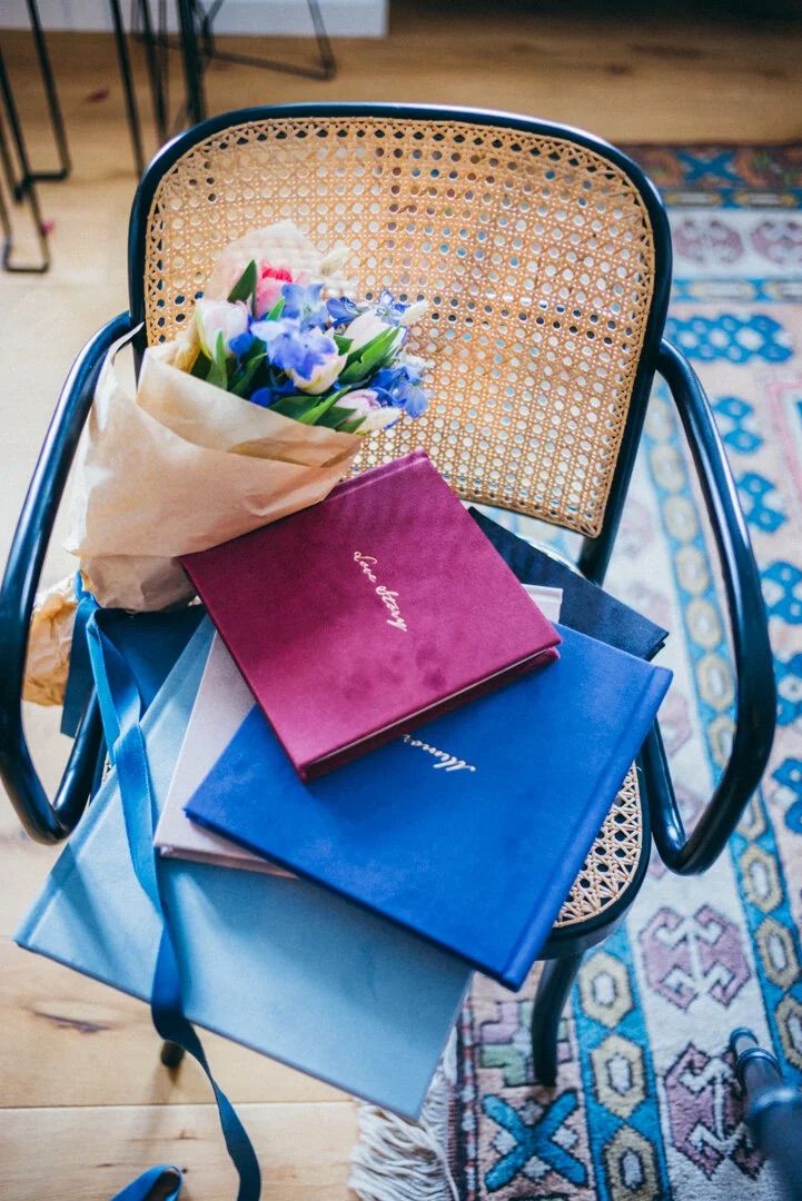 Elegant stack of velvet journals with a blue floral bouquet on a cane chair beside a colorful rug.