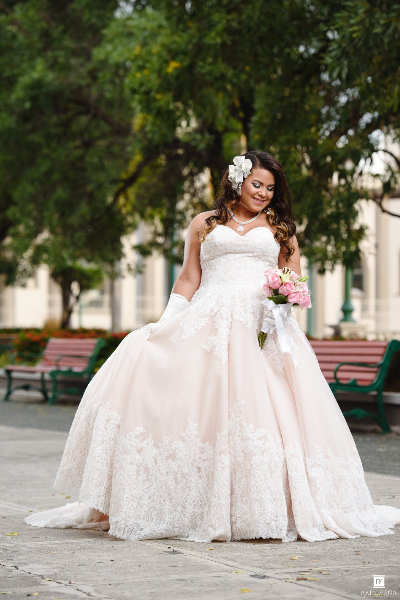 Bride in a strapless lace ball gown holding a pink bouquet, wearing a floral hair accessory outdoors.