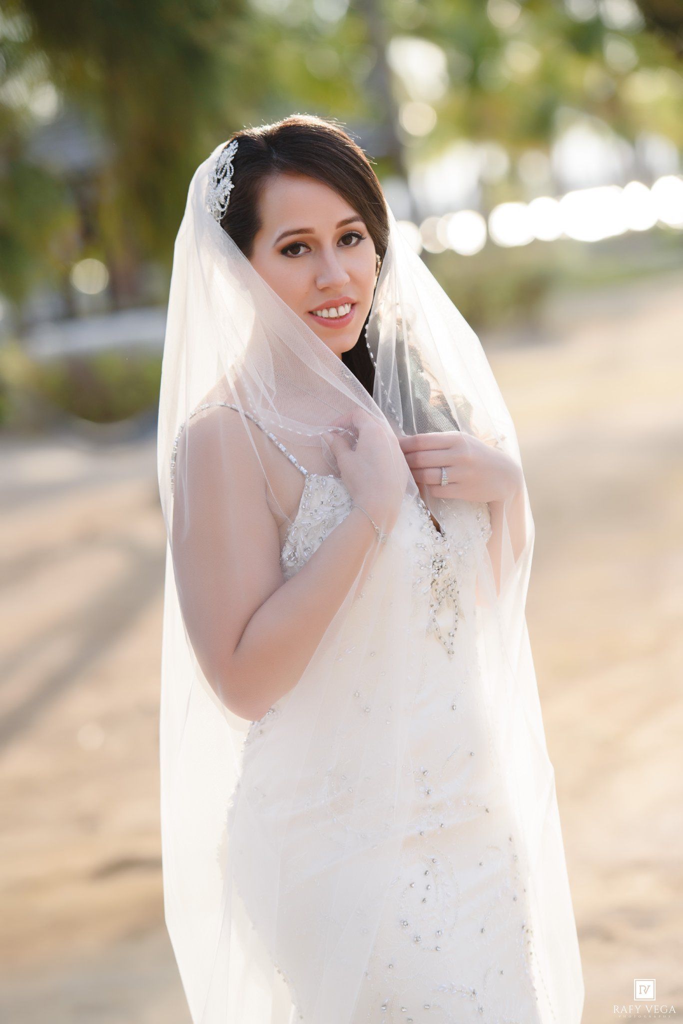 Smiling bride in beaded white gown and veil poses outdoors on sandy path with soft bokeh background.