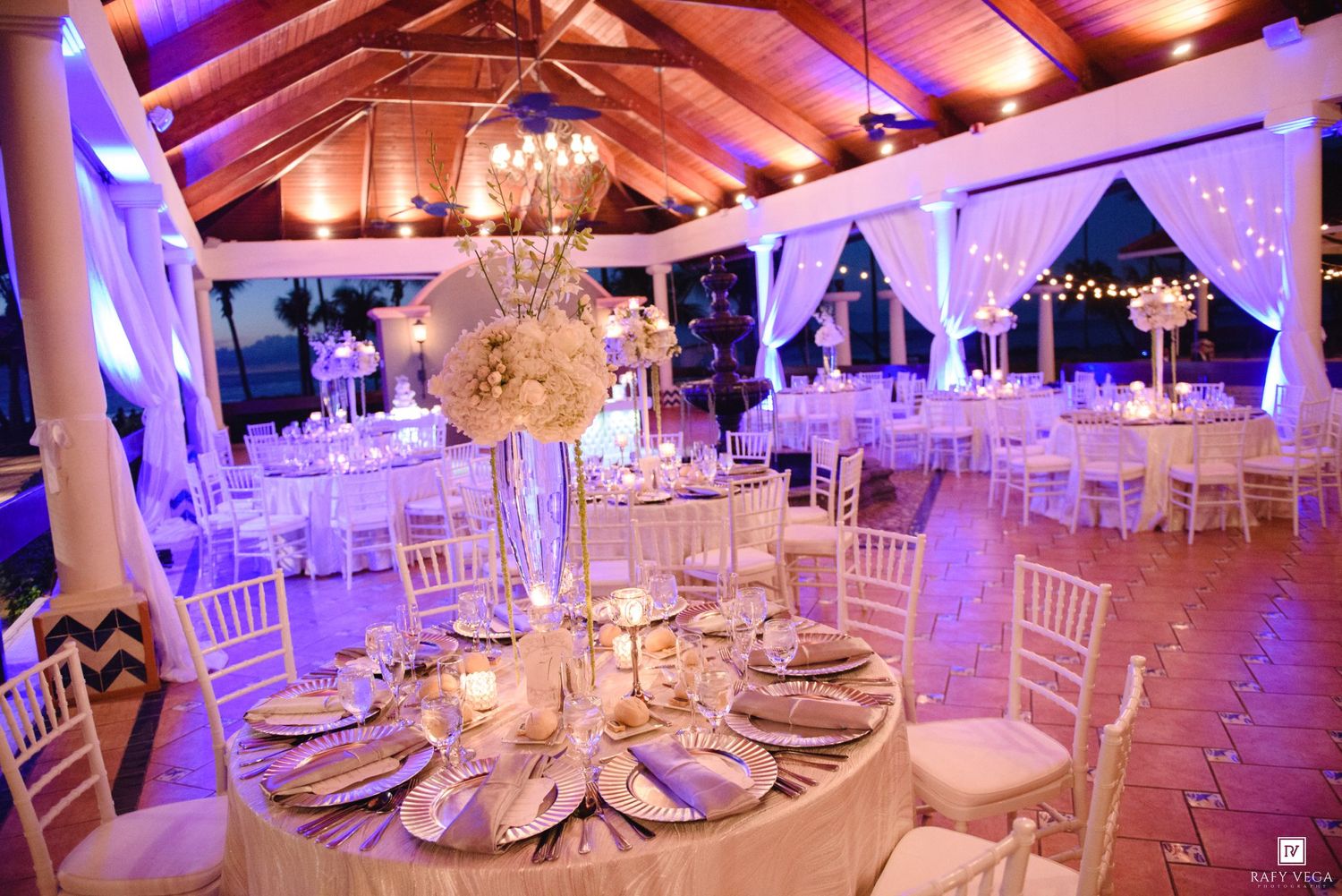 Elegant wedding reception venue with white floral centerpieces, chiavari chairs, candlelit tables, and purple ambient lighting.