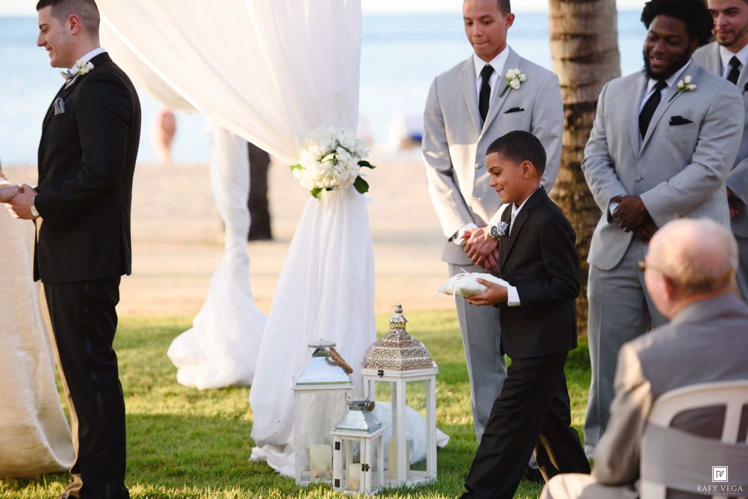 A young ring bearer in a black suit walks down the aisle at an elegant outdoor beach wedding ceremony.