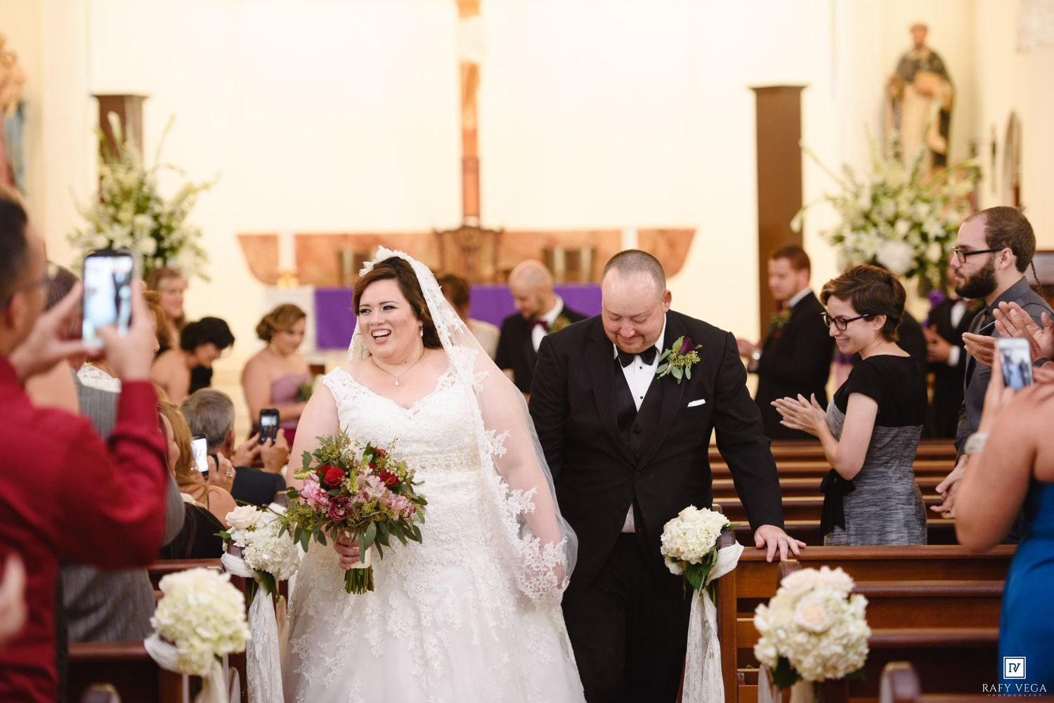 Bride and groom walk down church aisle after wedding ceremony, guests applauding, bride holding red and white floral bouquet.