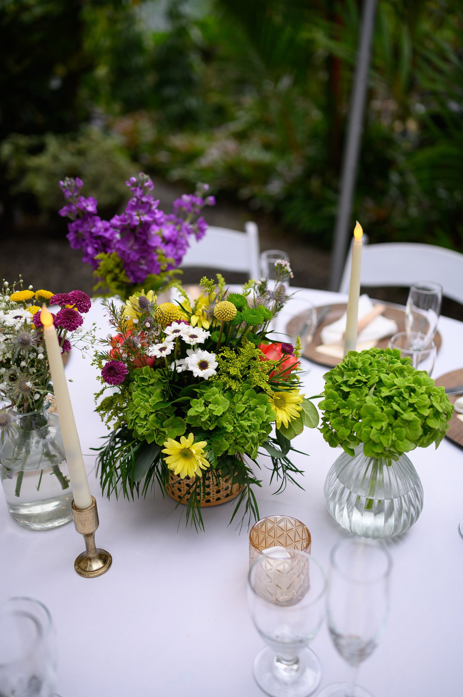 Colorful floral centerpiece with yellow, white, and red blooms, green hydrangeas, and taper candles on a white tablecloth.
