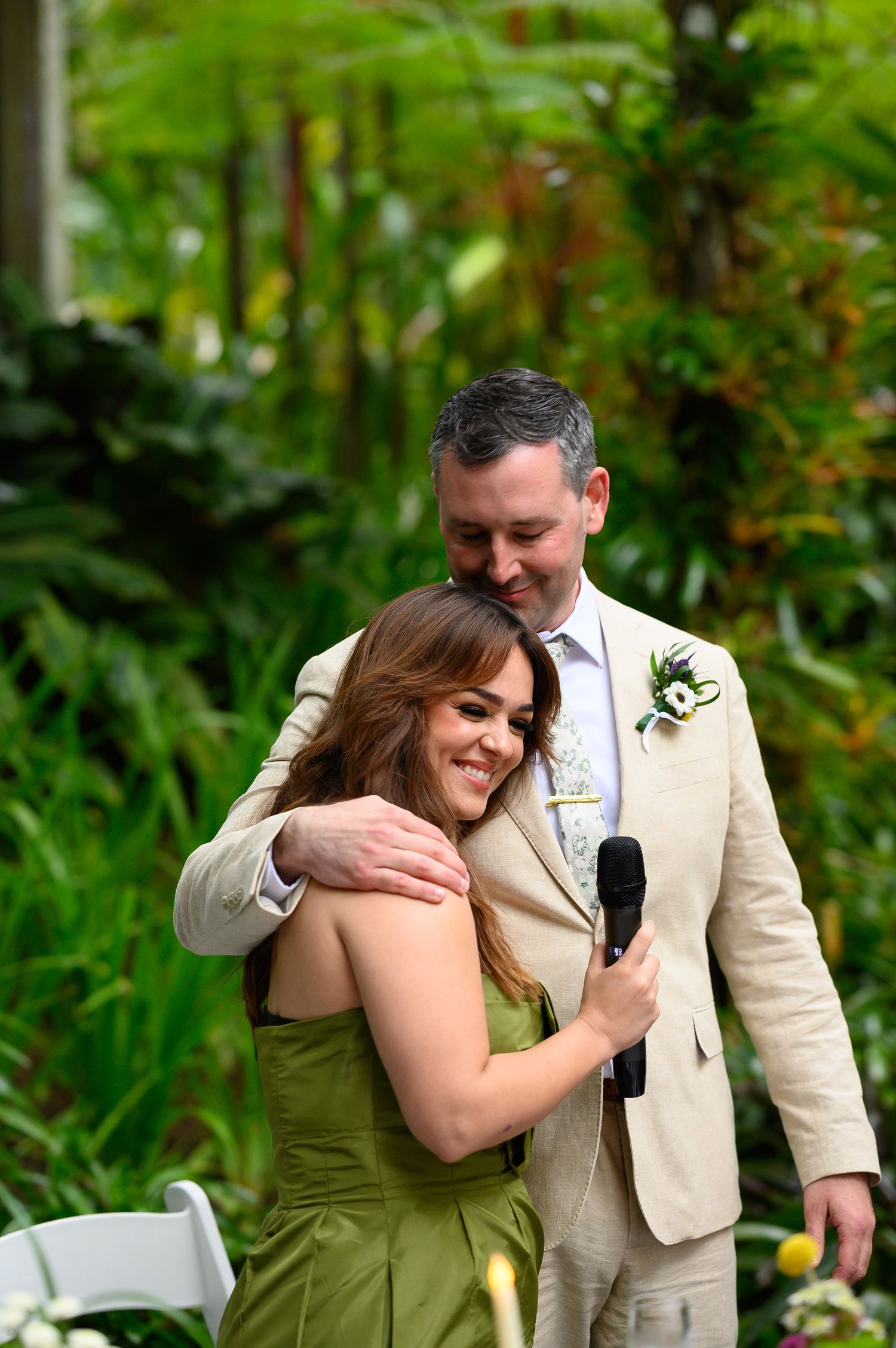 A smiling woman in a green dress holds a microphone while a man in a beige suit embraces her at an outdoor wedding ceremony.