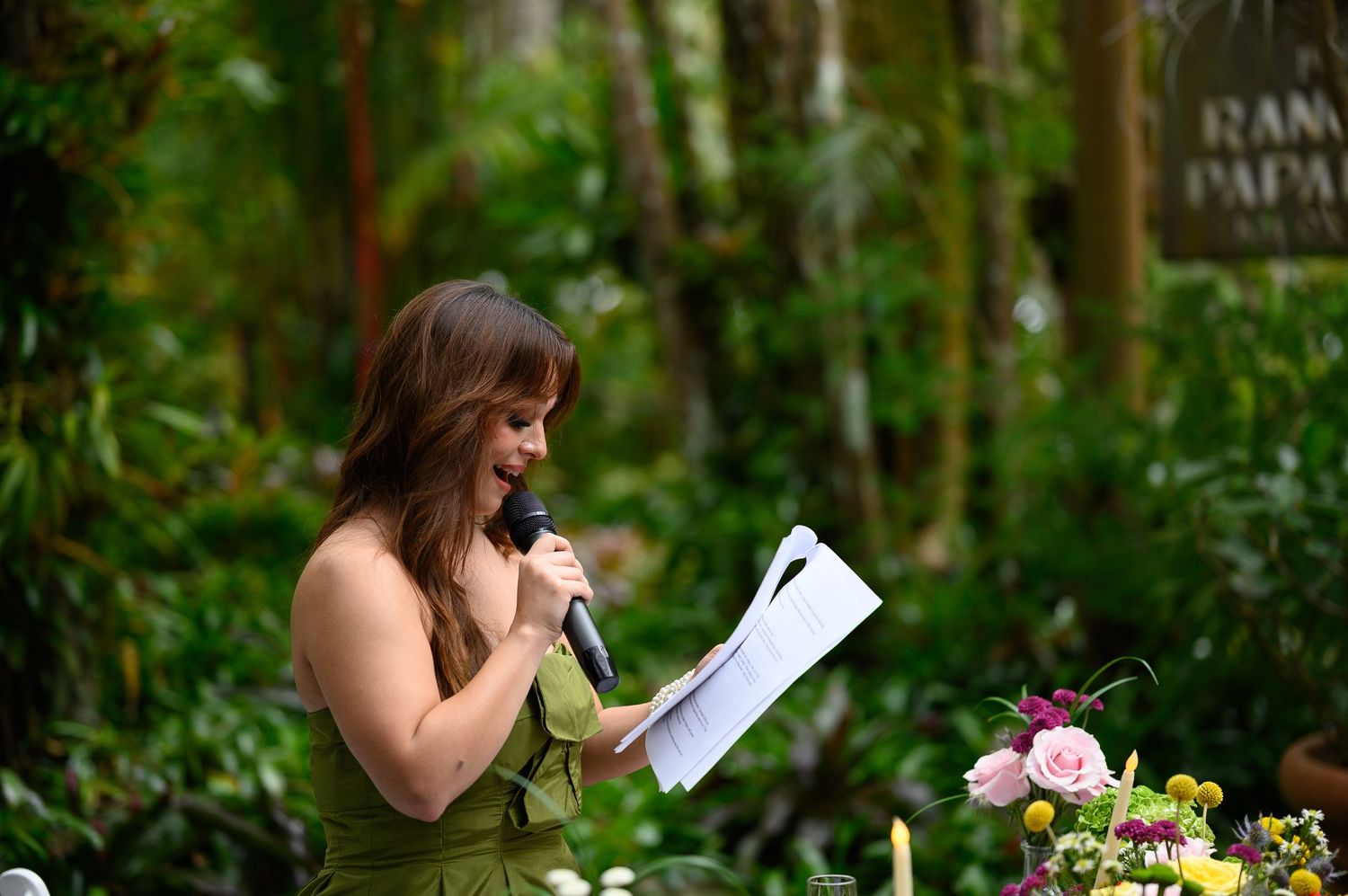 Woman in green dress speaks into microphone while holding papers at an outdoor garden event surrounded by lush tropical foliage.
