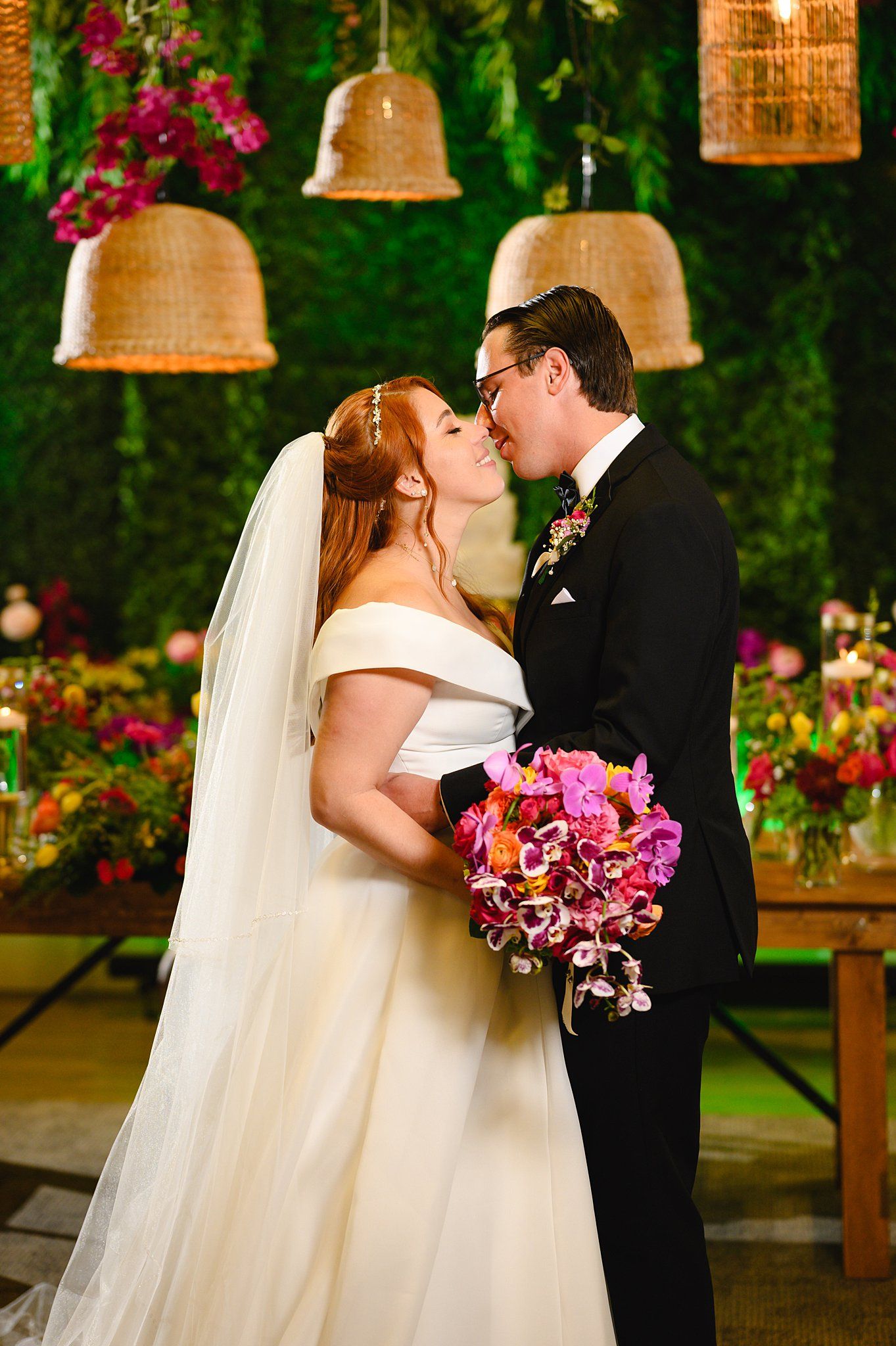 Bride and groom share first kiss at altar with lush green floral backdrop, rattan pendant lights, and vibrant pink bouquet.