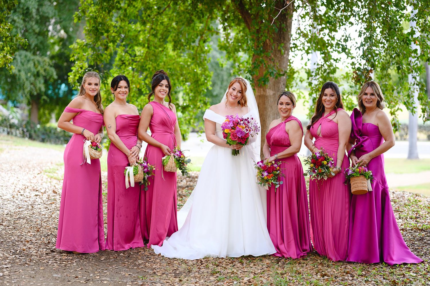 Bride in white gown poses with six bridesmaids in pink dresses holding bouquets outdoors under green trees.
