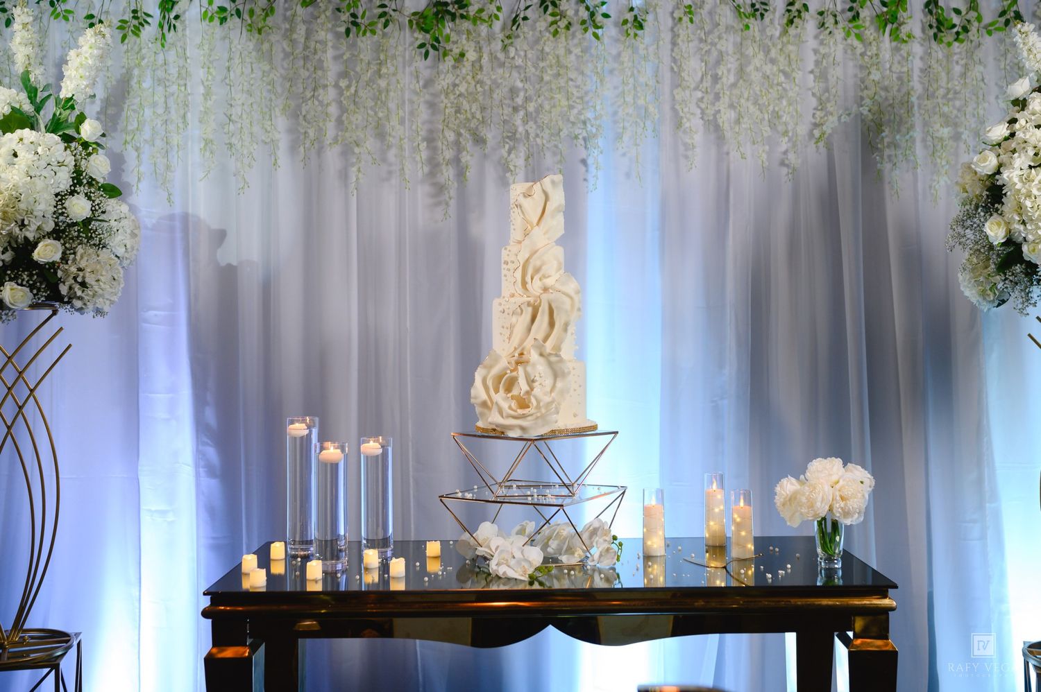 Elegant wedding cake display on wooden table with candles, white floral backdrop, and geometric stand at a reception venue.