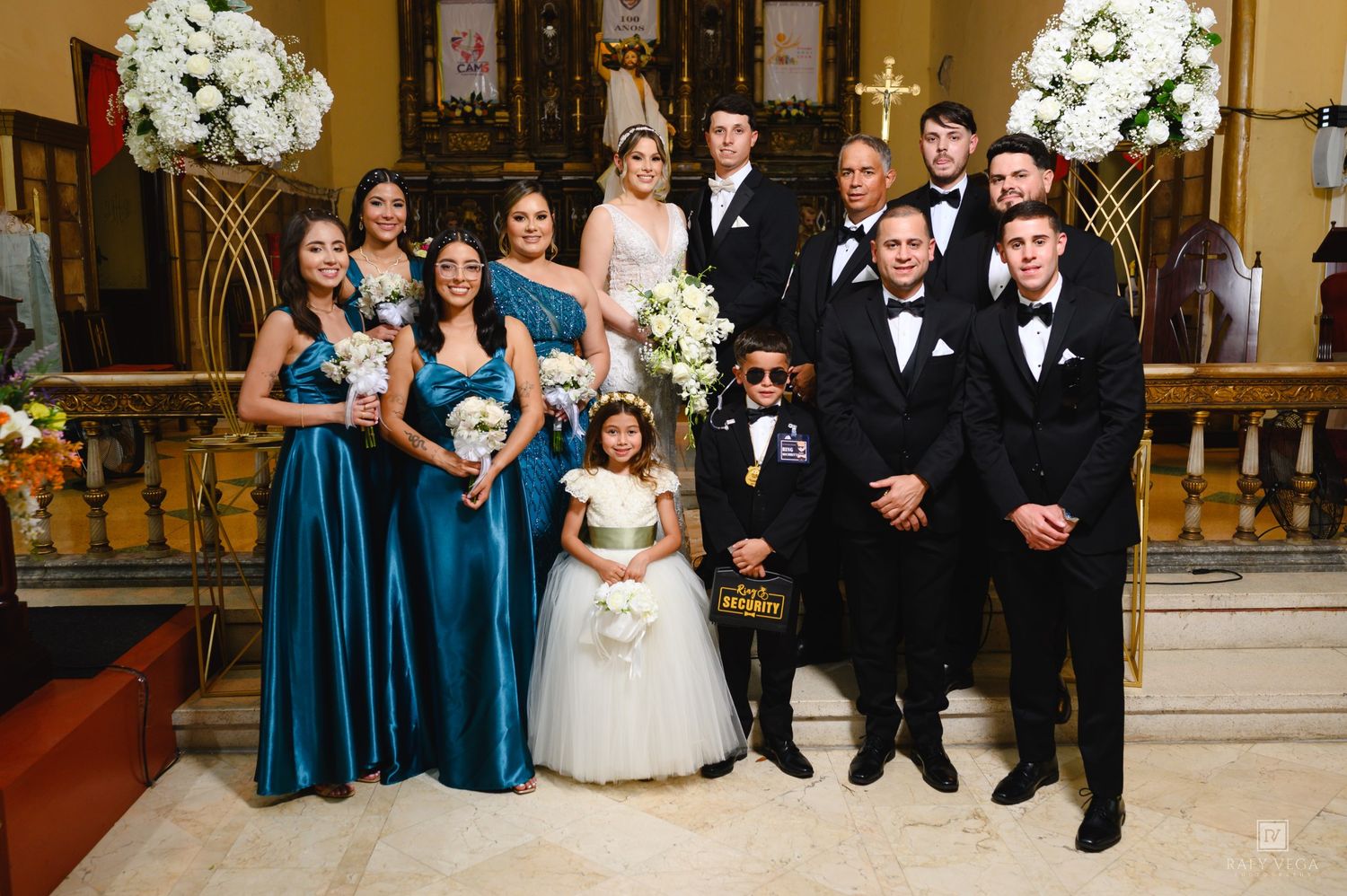 Wedding party poses in a church with bridesmaids in teal gowns, groomsmen in tuxedos, and flower girl in white dress.