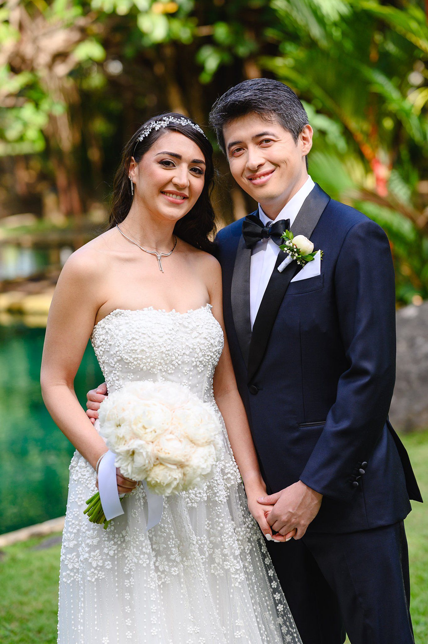 Bride in strapless pearl gown holding white bouquet stands with groom in navy tuxedo at outdoor garden wedding.