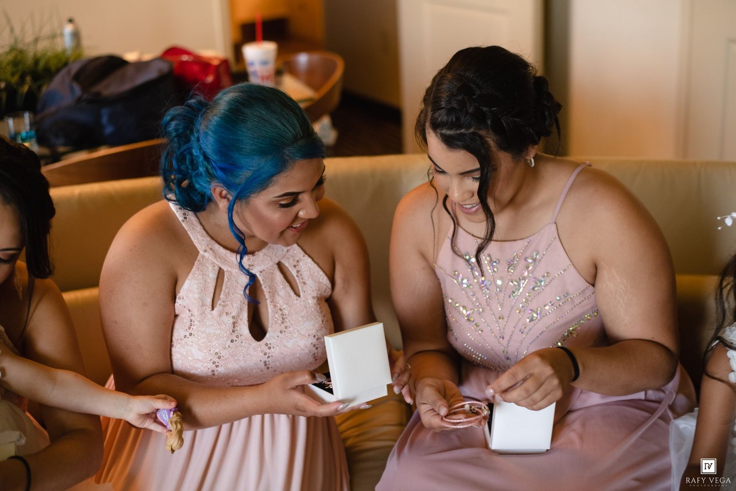 Two bridesmaids in blush pink dresses examine jewelry gifts, one with blue hair, during wedding preparations.