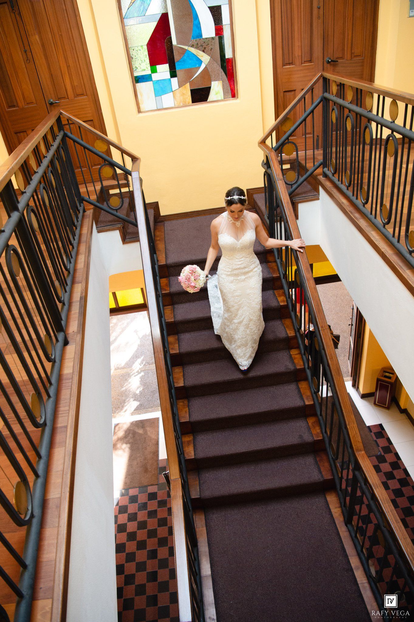 Bride in white gown holding pink bouquet descends elegant staircase with iron railings and colorful stained glass artwork.