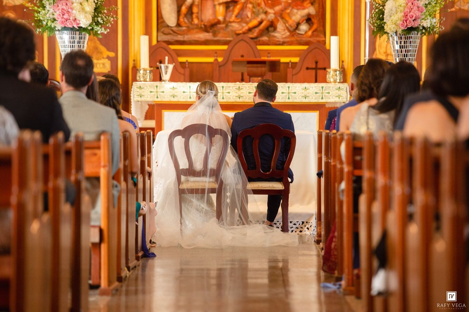 A bride and groom sit at the altar during a traditional church wedding ceremony, surrounded by guests in wooden pews.