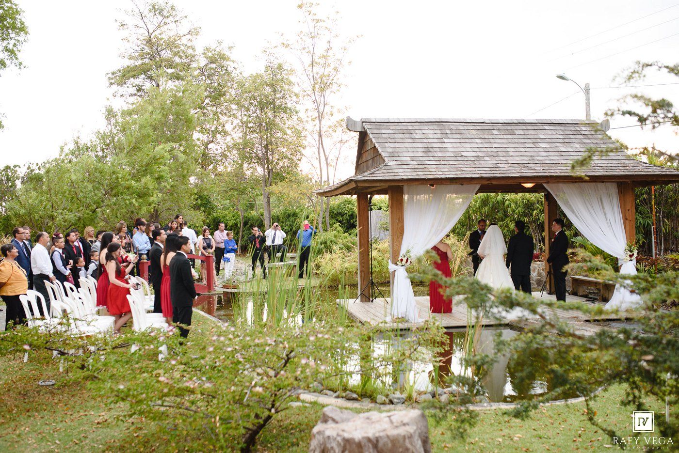 Outdoor wedding ceremony in a garden with guests seated along an aisle leading to a floral arch gazebo.