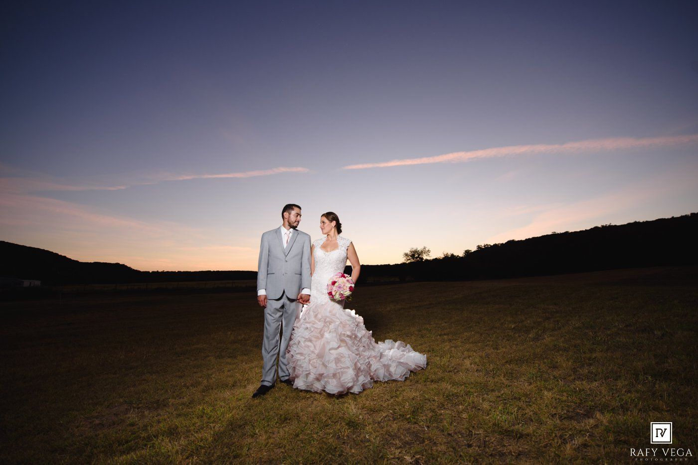 Bride and groom stand together in a field at sunset, bride wearing a ruffled wedding gown, groom in a gray suit.