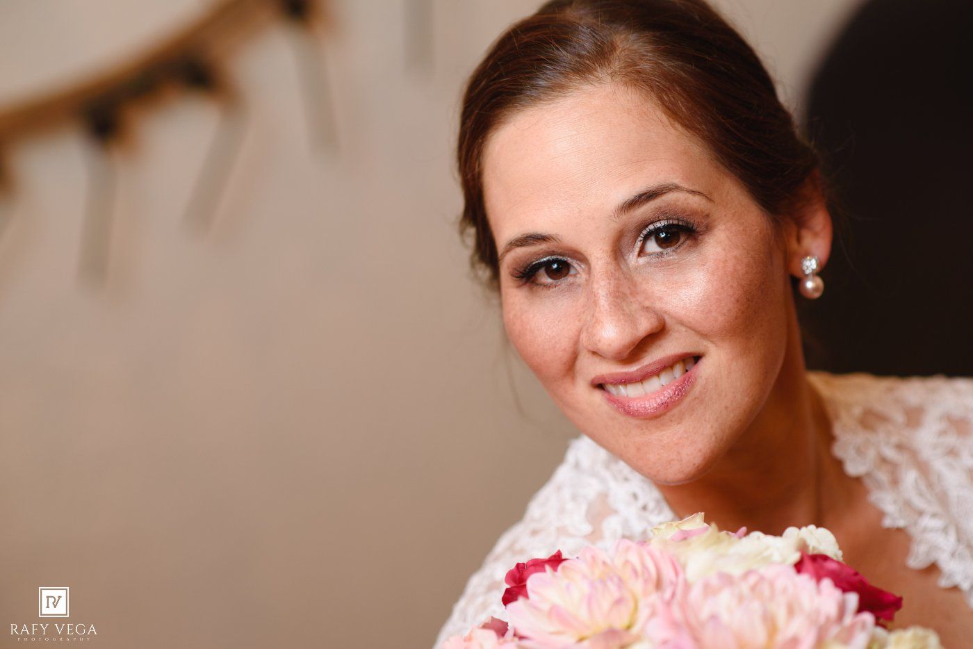 Smiling bride holding pink peony bouquet in lace wedding dress with pearl earrings on her wedding day.