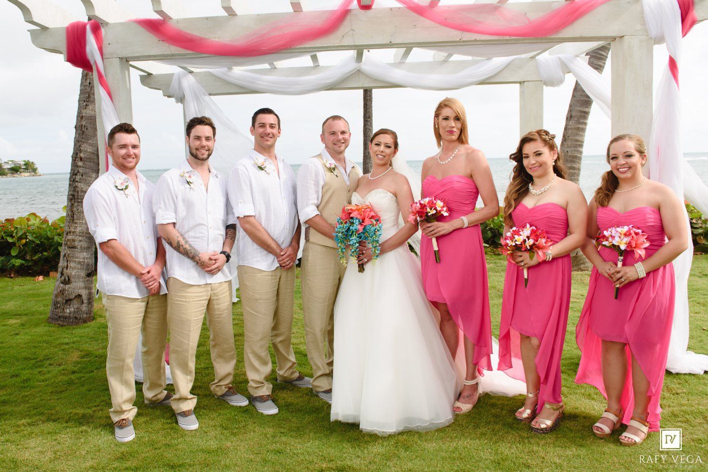 Wedding party posing outdoors in Hawaii, bridesmaids in pink dresses, groomsmen in khaki, bride and groom in center.