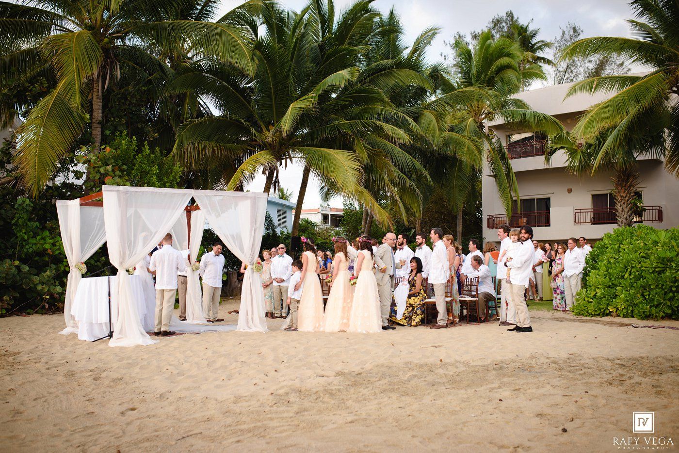 Beach wedding ceremony with guests gathered under a white canopy arch, surrounded by palm trees and a tropical resort backdrop.