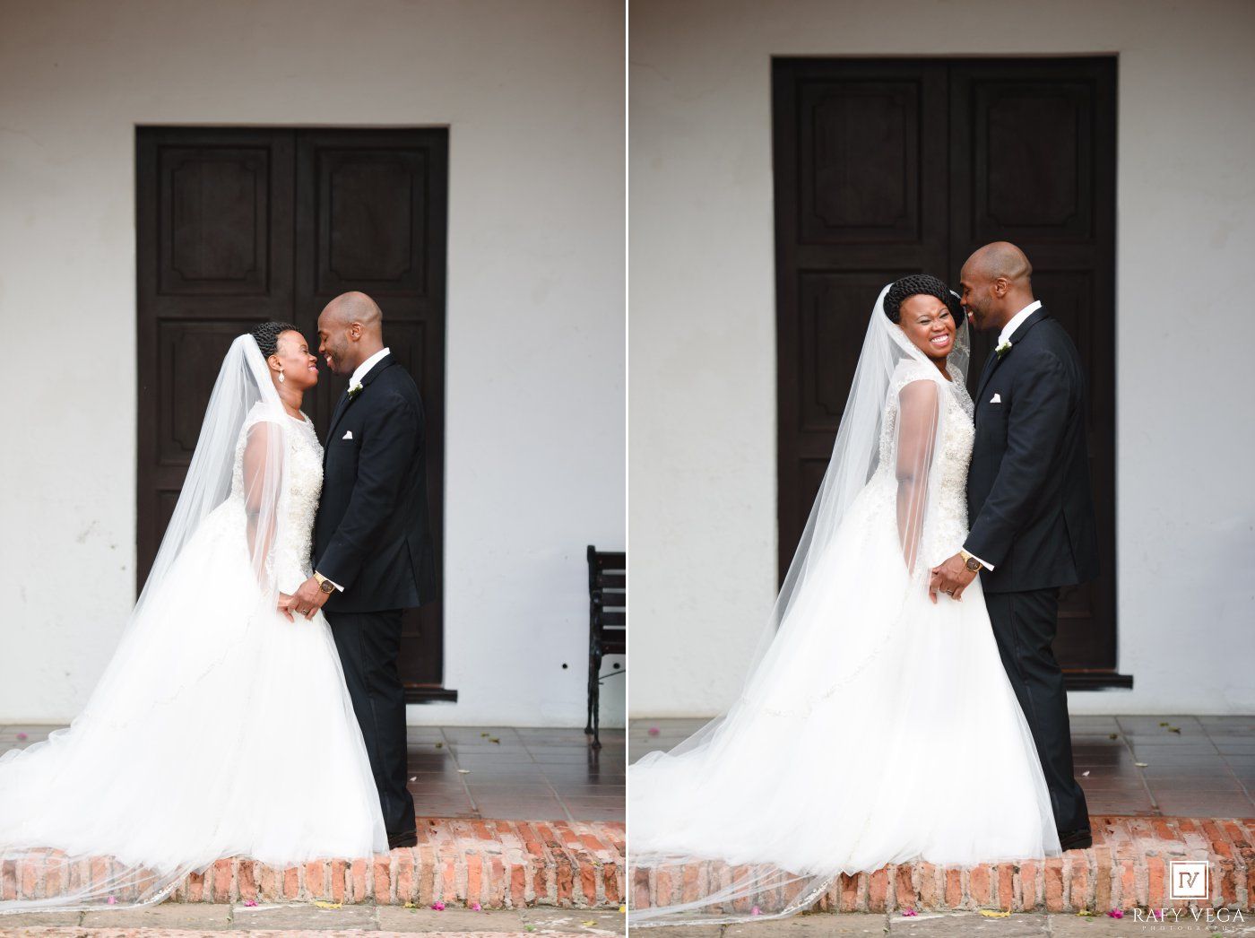 Wedding couple embracing before white building doors, bride in elegant ballgown with long veil and groom in black suit.