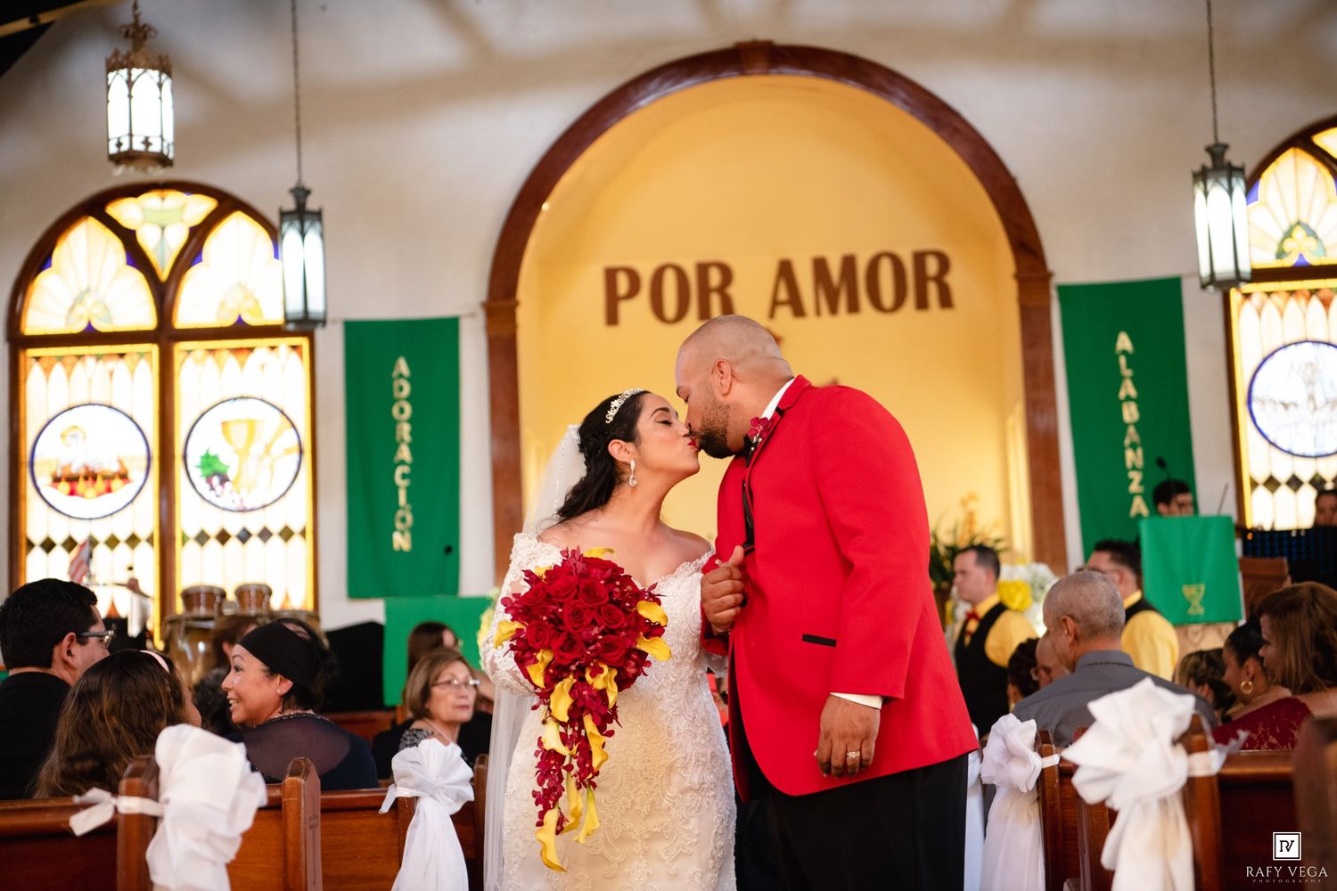 Newlywed couple sharing a kiss in a church aisle under a 'Por Amor' sign, bride holding red bouquet.